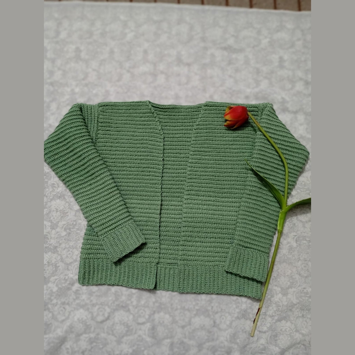 Green crochet rib stitch cardigan laid flat with a tulip