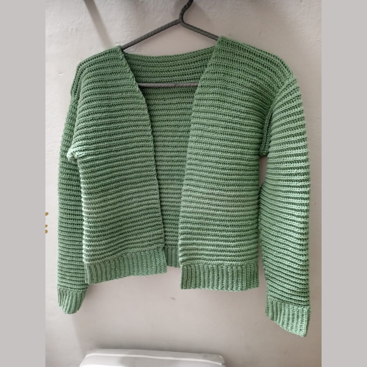Open-front green crochet ribbed cardigan hanging against a white wall